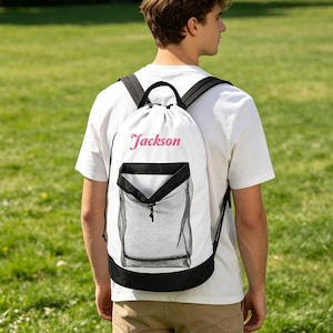 Puede incluir: Una mochila de cordón blanca con ribetes negros, correas negras para los hombros y un bolsillo frontal de malla negra. El nombre Jackson está bordado en letras cursivas rosas en la parte superior del cuerpo de tela blanca de la bolsa.