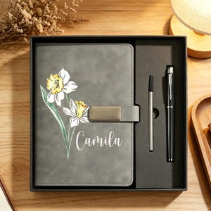 Op de afbeelding: Een grijs notitieboek met een illustratie van narcissen en de naam Camila in het wit gedrukt, voorzien van een metalen sluiting. Het notitieboek wordt gepresenteerd in een zwarte geschenkdoos met een zwarte balpen en een zilverkleurige vulling.