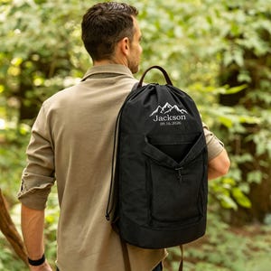 May include: A black drawstring backpack featuring a white mountain graphic, the name Jackson, and the date 09.16.2026 printed on the front. The bag includes a top handle, a front zippered pocket with a mesh overlay, and black cord shoulder straps.
