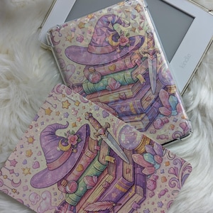 Könnte beinhalten: Ein Kindle E-Reader mit einer transparenten Hülle mit einer skurrilen Illustration. Das Design umfasst einen lila Hexenhut, Bücher, Kristalle und einen Dolch. Die Hülle und der E-Reader liegen auf einer flauschigen weißen Oberfläche.