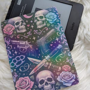 Könnte beinhalten: Ein schwarzer Kindle E-Reader mit einer dekorativen Hülle. Das Cover-Design zeigt Totenköpfe, Rosen, Waffen und ein Messer vor einem dunklen Hintergrund mit Regenbogenverlauf. Der Kindle ist ein beliebtes Lesegerät.