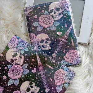 Könnte beinhalten: Ein Kindle E-Reader mit einer dekorativen Hülle mit einem Totenkopf-, Rosen- und Dolchmuster. Die Hülle hat einen dunklen Hintergrund mit rosa, lila und blauen floralen und gotischen Elementen. Das Design umfasst Herzen und Kugeln.