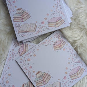 Könnte beinhalten: Ein Stapel quadratischer Notizblöcke mit einem skurrilen Design zum Thema Bücher. Jeder Notizblock zeigt Illustrationen von Büchern, Kaffeetassen, Brillen und floralen Akzenten in sanften Pastellfarben. Der Rand ist mit Sternen und Herzen verziert.