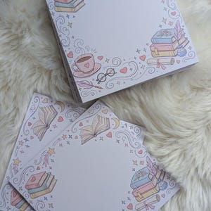 Könnte beinhalten: Quadratische Notizblöcke mit einem skurrilen Design zum Thema Bücher. Jeder Notizblock hat einen Rand aus Büchern, Herzen, Sternen und einer Tasse Tee. Die Farbpalette umfasst sanfte Rosa-, Blau- und Gelbtöne.