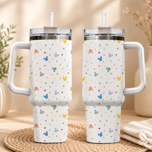 Pode incluir: Dois copos de a&ccedil;o inoxid&aacute;vel branco com al&ccedil;as e tampas transparentes com canudos. Os copos apresentam um padr&atilde;o repetitivo de pequenos pontos coloridos e min&uacute;sculas silhuetas de cabe&ccedil;as de rato em tons de azul, amarelo, laranja e cinza.