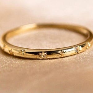 Puede incluir: Un anillo dorado con grabados en forma de estrella y pequeñas piedras preciosas. El anillo se encuentra sobre un fondo neutro, resaltando su acabado pulido y su delicado diseño. El anillo mide aproximadamente 0,2 cm de ancho.