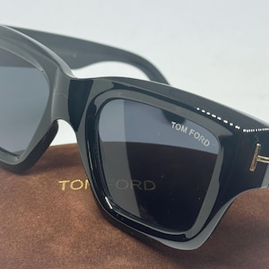 May include: Black sunglasses with thick frames and dark lenses. The right lens features the white text TOM FORD. A gold-toned T-shaped metal accent is visible on the temple hinge. The sunglasses are resting on a brown velvet-textured case.