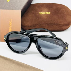 Peut inclure: Lunettes de soleil style aviateur noires avec des accents dorés en forme de T sur les charnières. Les verres sont sombres et la monture porte le logo Tom Ford sur le coin supérieur. Inclut un étui en velours marron, une boîte, une carte et un chiffon.