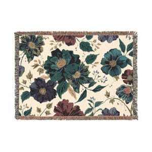 Könnte beinhalten: Eine rechteckige gewebte Wurkdecke mit floralem Muster. Das Design zeigt große Blumen in Teal-, Lila- und Burgundertönen auf cremefarbenem Hintergrund. Die Decke hat einen Fransenrand.