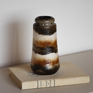 Puede incluir: Un jarrón de cerámica vintage con un patrón a rayas en tonos marrón, blanco y naranja. El jarrón es cilíndrico con una base ligeramente ensanchada y está sobre un libro con texto descolorido. El jarrón mide unos 20 cm de alto.