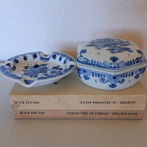 May include: A white ceramic ashtray and a matching lidded box, both decorated with blue floral patterns. The ashtray has a circular shape with indentations, while the box is hexagonal. They are placed on two stacked books with visible text.