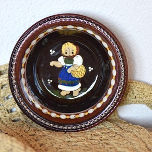 Op de afbeelding: Een ronde, bruine keramische schaal met een geschilderd volkskunstontwerp. De schaal heeft een centraal beeld van een vrouw in traditionele kleding die een mand draagt. De rand is versierd met witte en gele accenten.