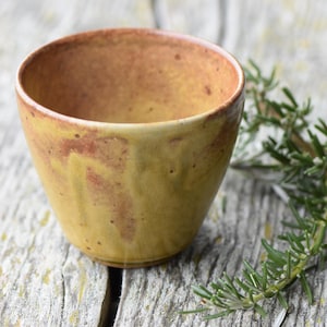 May include: A handmade ceramic cup with a yellow and brown glaze. The cup has a rounded shape and sits on a wooden surface. A sprig of rosemary is next to the cup.