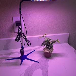 Op de afbeelding: Een zwarte LED-groeilamp met een blauwe driepootvoet verlicht een kleine plant in een pot. De lamp heeft rode, blauwe en witte LED's. De plant staat in een kleine bruine pot. De achtergrond is een wit aanrecht en een getextureerde muur.