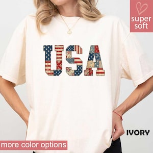 May include: Ivory-colored t-shirt with the word "USA" in a patchwork design featuring red, white, and blue patterns. The shirt has short sleeves and a relaxed fit. The words "super soft" and "more color options" are also visible.