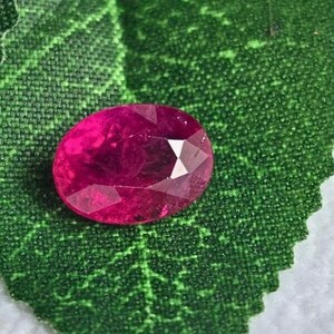 Könnte beinhalten: Ein facettierter, oval geformter Rubin-Edelstein. Der Rubin hat eine tiefe, lebendige rote Farbe mit mehreren Facetten, die das Licht einfangen. Er liegt auf einem strukturierten, grünen Blatt und erzeugt einen auffälligen Kontrast.