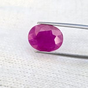 Peut inclure: Une pierre précieuse de rubis ovale et facettée, tenue par une pince. Le rubis est d'un magenta profond, avec de multiples facettes captant la lumière. Le fond est un tissu blanc texturé.