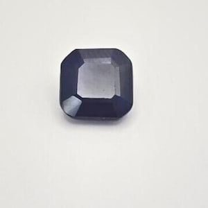 May include: A faceted, dark blue gemstone with an emerald cut. The gem is square with beveled edges and reflects light, showcasing its facets. The stone is set against a white background.