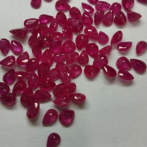 May include: A scattering of faceted, teardrop-shaped rubies in various sizes. The gemstones are a deep, vibrant red colour and are spread across a light-coloured surface. The gems have a polished, reflective surface.