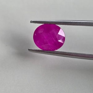 Może przedstawiać: Fasetowany, owalny kamień szlachetny rubinu trzymany przez srebrną pęsetę. Rubin ma żywy, różowo-czerwony odcień, z wieloma fasetami odbijającymi światło. Pęseta jest umieszczona na gładkim, jasnym tle.
