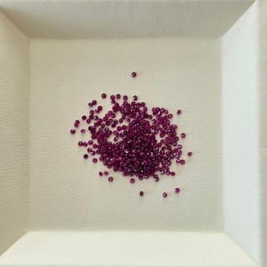 Pode incluir: Um conjunto de pequenas pedras preciosas facetadas, vermelho escuro, provavelmente rubis, está espalhado sobre uma superfície branca texturizada. As pedras preciosas estão agrupadas no centro de uma bandeja quadrada, com algumas pedras espalhadas nas bordas.