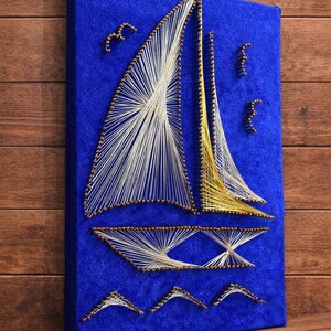May include: A sailboat string art piece on a blue canvas. The boat features white and yellow sails, with brown accents and a blue background. The artwork includes small bird-like shapes. The piece is mounted on a wooden surface.