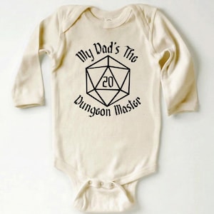 May include: Cream-colored baby bodysuit with long sleeves. The front features the text "My Dad's The Dungeon Master" surrounding a black D20 die graphic. The bodysuit has snap closures at the bottom.