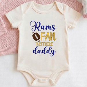 May include: Cream-colored baby onesie with the text "Rams FAN just like daddy" in blue and gold. A brown football graphic is in the center. A pink sweater and a white unicorn plush toy are in the background.