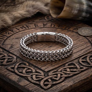 Op de afbeelding: Een zilverkleurige, gevlochten kettingarmband met een magnetische sluiting. De armband wordt getoond op een houten oppervlak met gesneden Keltische knoopontwerpen. Een vacht en een hoorn zijn op de achtergrond.