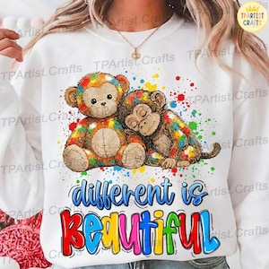 May include: White sweatshirt featuring a colorful graphic of two monkeys, one resembling a teddy bear. The monkeys are surrounded by paint splatters and the words "different is beautiful" in a rainbow font.
