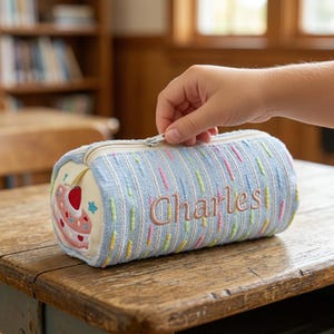 May include: A light blue pencil case with the name "Charles" embroidered in a light brown thread. The cylindrical case has a zipper closure and a colorful vertical stripe pattern. One end features a cake design.