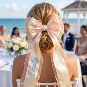 Può includere: Un grande fiocco per capelli color crema e rosa, indossato sulla nuca di una persona. Il fiocco presenta lunghi nastri con il nome "Emily" ricamato in azzurro. Lo sfondo mostra un matrimonio sulla spiaggia.