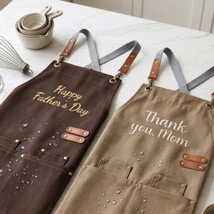 May include: Two aprons with cross-back straps, one dark brown with "Happy Father's Day" embroidered in gold, and one tan with "Thank you, Mom" embroidered in white. Both feature leather-like accents, metal rivets, and multiple front pockets.