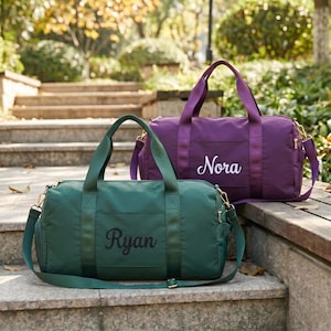 Könnte beinhalten: Zwei grüne und lila Sporttaschen stehen auf Steinstufen. Auf der grünen Tasche ist der Name "Ryan" aufgestickt. Auf der lila Tasche ist der Name "Nora" aufgestickt. Beide Taschen haben Griffe und Schultergurte.