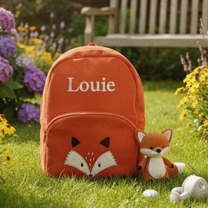 Może przedstawiać: Pomarańczowy plecak z motywem liska i imieniem "Louie" wyhaftowanym na biało. Pluszowa zabawka w kształcie lisa i mała konewka leżą na trawie. Plecak ma przednią kieszeń i regulowane paski.