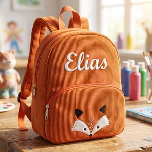 Puede incluir: Mochila de pana naranja con el nombre "Elias" bordado en blanco. La parte delantera presenta un diseño de cara de zorro. Incluye asa superior, correas ajustables y un bolsillo frontal con cremallera.