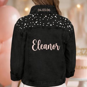 May include: Black denim jacket adorned with pearl studs on the shoulders. The name "Eleanor" is written in pink script on the back. The date "04.03.06" is above the collar. A fashionable personalized jacket.