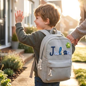 May include: A light grey corduroy backpack featuring the name Julian embroidered in blue and white script lettering on the front pocket. A small green and yellow crocheted flower is attached to the upper right corner. The bag has white rope zipper pulls.