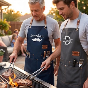 Può includere: Due grembiuli con cinghie regolabili in pelle marrone con clip in metallo, tasche multiple e rivetti in metallo. Il grembiule blu navy mostra il nome Nathan con un grafico a baffi, quello grigio antracite il nome Daniel con un monogramma.