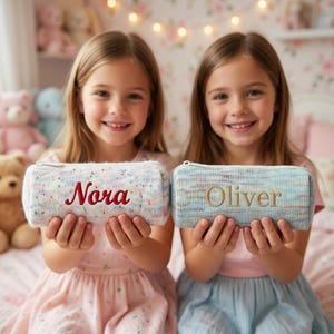 May include: Two pencil cases, one white with red embroidered text "Nora", the other blue with gold embroidered text "Oliver". Both cases have a zipper closure and are held by young girls.