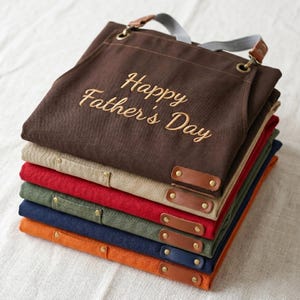 May include: A stack of folded aprons in brown, beige, red, green, blue, and orange. The top brown apron features the embroidered text Happy Father's Day in a light script font. Each apron includes brown leather accents and metallic rivets.