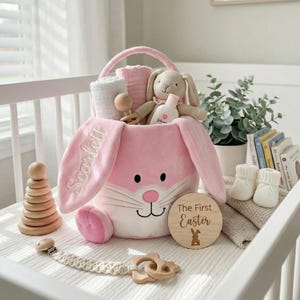 May include: A pink bunny-shaped Easter basket filled with baby items. The basket has embroidered lettering and a handle. Items include a stuffed bunny, baby lotion, a wooden rattle, and a wooden disc that says "The First Easter".