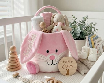 May include: A pink bunny-shaped Easter basket filled with baby items. The basket has embroidered lettering and a handle. Items include a stuffed bunny, baby lotion, a wooden rattle, and a wooden disc that says "The First Easter".