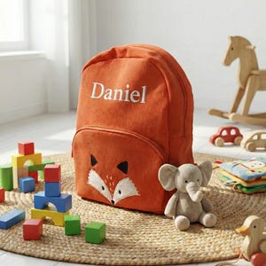 May include: An orange corduroy backpack embroidered with the name "Daniel". The backpack has a fox face design and a front pocket. Toys, including a plush elephant and wooden blocks, are scattered around it.