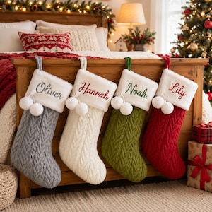 May include: Four cable-knit Christmas stockings hanging in a row. Each stocking features a white faux fur cuff with two white pom-poms and embroidered names: Oliver in gray, Hannah in red, Noah in green, and Lily in red. The stockings are gray, white, olive green, and red.