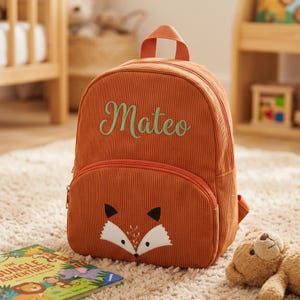 Puede incluir: Una mochila de pana naranja con un diseño de cara de zorro en el bolsillo delantero. El nombre "Mateo" está bordado en verde en la parte delantera. La mochila tiene un asa superior y correas ajustables.