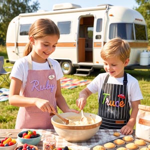 Peut inclure: Deux enfants portant des tabliers rayés, mélangeant une pâte à gâteau à l'extérieur. L'un est rouge et blanc avec le nom "Ruby", l'autre est noir et blanc avec le nom "Trude" et un dessin de voiture. Une caravane vintage est en arrière-plan.