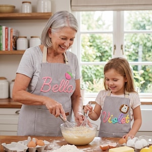 Op de afbeelding: Een vrouw en een kind dragen gestreepte schorten met de namen "Bella" en "Violet" in een keuken. De schorten hebben een streepjespatroon met een aardbei- en egelontwerp. Ze mengen ingrediënten in een glazen kom met gardes.