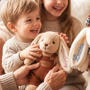 Könnte beinhalten: Ein beiges Plüschkaninchen in brauner Cord-Latzhose mit einem Lederaufnäher mit der Aufschrift MUSITU. Ein Ohr ist mit dem Wort Birthday in rosa Garn bestickt, während ein weiteres Plüschtier in der Nähe den Namen Lucas in Blau trägt.