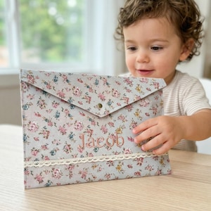 Puede incluir: Un portadocumentos con estampado floral con el nombre "Jacob" bordado en rosa. La tela azul claro presenta un estampado de flores rosas, azules y amarillas. Un ribete de encaje adorna el borde inferior. El portadocumentos tiene un cierre de solapa con un broche.